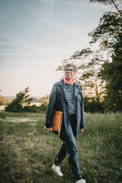 Carmen Müller im Grünen bei Sonnenuntergang, trägt eine lange Jacke, Brille und trägt eine braune Rednermappe, im Freien in der Natur.