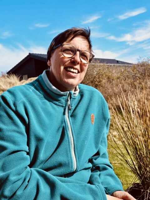Carmen Müller im blauen Fleece vor ländlicher Kulisse mit Himmel und Häusern, lächelnd, bei Abschied mit Weitblick, Natur, Outdoor, positive Stimmung, Carmen Müller.